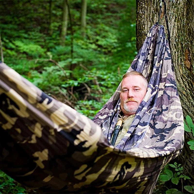 comfortable looking mans face pokes out of a camouflage hammock in the forest